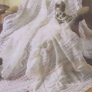 May include: A white crocheted baby blanket with a delicate lace pattern. The blanket is draped over a chair and has a small white dog toy on top.