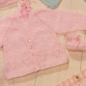 Puede incluir: Un cárdigan de punto rosa para bebé con cierre de botones y detalles en forma de corazón. El cárdigan está hecho con un hilo suave y esponjoso.