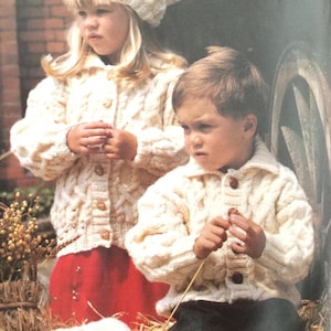 May include: Two children wearing white knitted cardigans with button closures. The girl is wearing a red skirt and a white hat. The boy is wearing black pants.