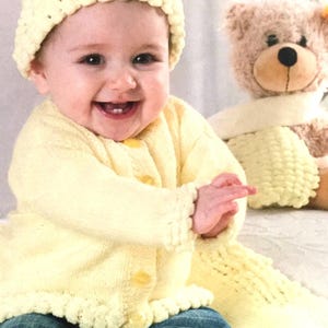 May include: A smiling baby wearing a pale yellow knitted hat and cardigan with matching scarf. The baby is wearing blue jeans and is sitting next to a teddy bear.