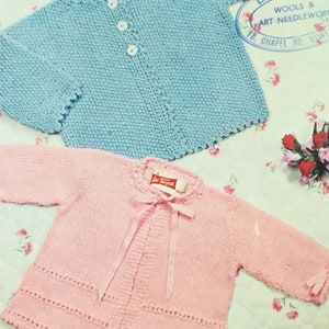 May include: Two knitted baby sweaters, one light blue and one pink, with button closures and ribbon ties. The sweaters are laid out on a floral patterned fabric.