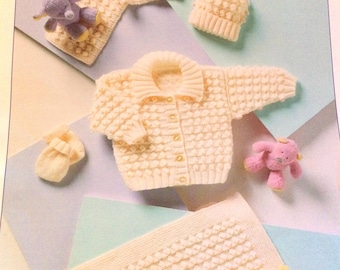 May include: A set of knitted baby clothes in a cream color, including a cardigan, a hat, and a pair of booties. The cardigan has a button closure and a collar. The hat is a simple beanie style. The booties are small and have a ruffled edge.