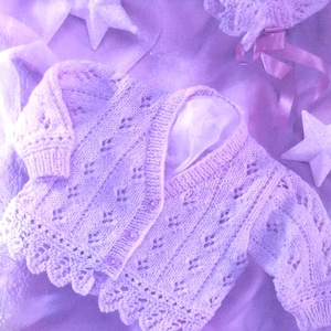 Puede incluir: Un delicado cárdigan de bebé de punto de color lila claro con un patrón de encaje. El cárdigan tiene un cierre de botón y un borde festoneado.