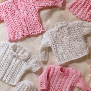 May include: Four knitted baby sweaters in pink and white. Two sweaters are white with a lacy pattern, one is pink with a lacy pattern, and one is pink with a ribbed pattern.