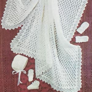 May include: A white crocheted baby blanket with a delicate lace pattern. The blanket is laid out on a red patterned surface with a white crocheted bonnet and booties.