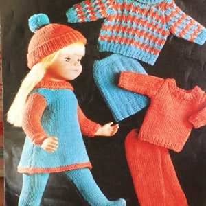 May include: A blue and red knitted doll outfit with a matching hat. The outfit includes a dress, leggings, and a sweater. The hat has a pom-pom on top.