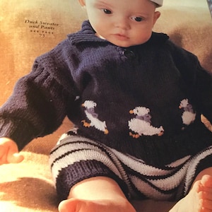 May include: A baby wearing a blue knitted sweater with a duck design and matching striped trousers. The sweater has a white collar and the text "Duck Sweater and Pants See Page 75" is printed on the sweater.