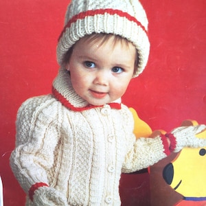 May include: A white knitted baby cardigan with red trim and buttons. The cardigan has a matching hat with a white pom-pom on top. The baby is wearing the cardigan and hat and is looking at the camera.