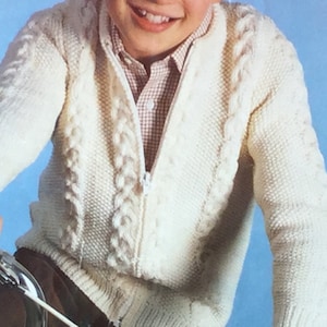 May include: A white knitted cardigan with a zipper closure and a cable knit pattern. The cardigan is worn by a child.