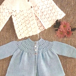 May include: Two knitted baby cardigans. One is white with a diamond pattern, and the other is light blue with a decorative neckline. Both have button closures. Pink flowers are next to the white cardigan.