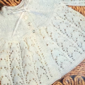 May include: A white crocheted baby cardigan with a button fastening and a delicate lace pattern.