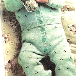 May include: A baby in a light green knitted outfit, comprising a collared cardigan and trousers, with small decorative holes. The baby holds a floral patterned soft toy.