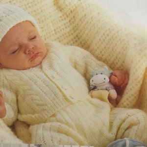 May include: A baby wearing a white knitted hat and a white knitted outfit with a cable knit pattern. The baby is holding a stuffed animal in the shape of a hippopotamus.