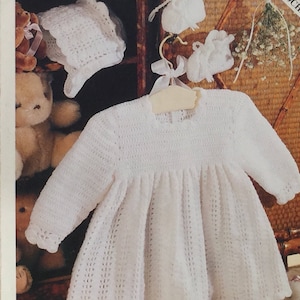 May include: White crocheted baby dress with long sleeves and a gathered skirt. The dress is hanging on a wooden hanger. There are two white crocheted hats and two pairs of white crocheted booties in the background.