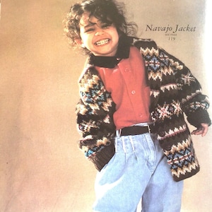 Könnte beinhalten: Ein junges Kind trägt einen braunen, weißen und blauen gemusterten Pullover mit einem roten Hemd mit Kragen darunter. Der Pullover hat ein Navajo Jacket-Etikett mit dem Text "Siehe Seite 119". Das Kind trägt auch blaue Jeans und einen schwarzen Gürtel.