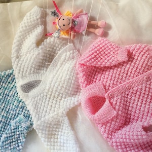 Puede incluir: Tres jerséis de punto para bebé en azul, blanco y rosa. Los jerséis tienen un patrón de punto texturizado y están dispuestos sobre un fondo blanco.