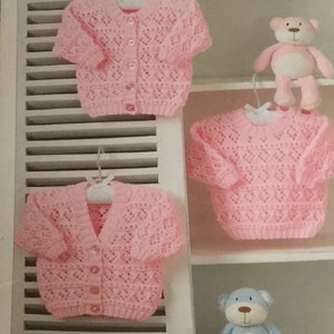 May include: Two pink knitted baby cardigans with button closures hanging on a white shelf. A pink teddy bear is on the shelf above the cardigans.
