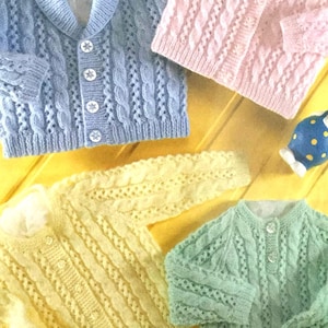 May include: An assortment of cable-knit baby cardigans in pastel colors: light blue, pink, yellow, and mint green. Each sweater features a button-up front and a classic design. A small ceramic bunny figurine is also present.
