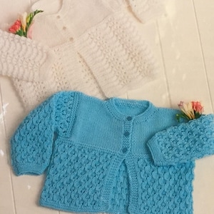 May include: Two knitted baby cardigans. One is white, the other is light blue. Both have button closures and textured sleeves and body. The cardigans are displayed on a light-coloured surface, with flowers tucked into the sleeves.