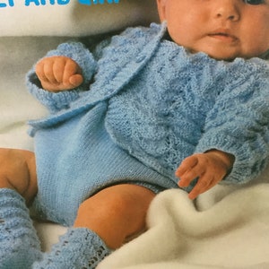 May include: A baby wearing a blue knitted sweater and matching leg warmers. The sweater has a cable knit pattern and the leg warmers have a lacy pattern. The baby is lying on a white blanket.