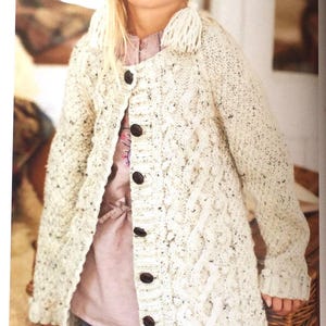 May include: A white cable knit cardigan with brown buttons. The cardigan has a loose fit and is worn by a child. The child is also wearing a white knit hat with a tassel.