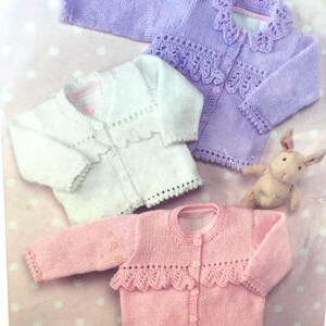 Puede incluir: Tres cárdigans de punto para bebé en blanco, rosa y lavanda con ribetes de ganchillo alrededor del cuello y los puños.