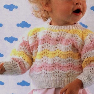 Könnte beinhalten: Ein gestrickter Pullover mit weißen, rosa und gelben Streifen und einem Lochmuster. Der Pullover hat einen runden Ausschnitt und lange Ärmel.