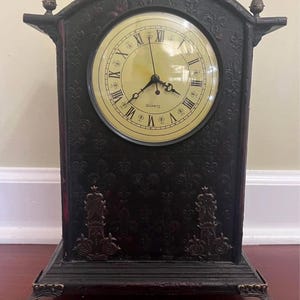 Puede incluir: Un reloj de sobremesa negro con esfera color crema, números romanos negros y la palabra QUARTZ impresa en negro. El reloj presenta una parte superior arqueada, remates metálicos decorativos, patrones en relieve y cuatro patas metálicas curvas.