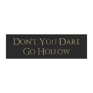 May include: A rectangular black sign featuring the text "DON'T YOU DARE GO HOLLOW" in a distressed, light-colored serif font. The lettering has a weathered appearance, contrasting against the solid dark background of the horizontal sign.