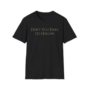 May include: A black short-sleeved t-shirt featuring the text "DON'T YOU DARE GO HOLLOW" printed in a weathered, metallic-toned font across the chest. The garment has a standard crew neckline and a straight hemline, constructed from solid black fabric.