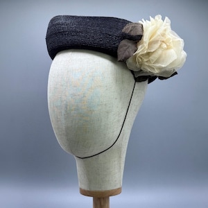 May include: A black pillbox hat featuring a textured, woven surface. The hat is adorned on the right side with a large, layered cream-coloured fabric flower and dark brown fabric leaves, secured with a thin black elastic chin strap.