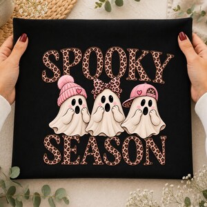 Puede incluir: Una camiseta negra con el texto SPOOKY SEASON en un patrón de estampado de leopardo marrón. Debajo, tres fantasmas blancos con expresiones de sorpresa llevan un gorro rosa con pompón, un lazo de leopardo y una gorra de béisbol rosa.