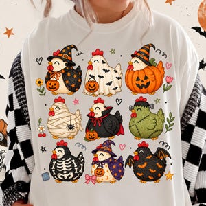 Op de afbeelding: Een wit sweatshirt met ronde hals en een grafische print van negen cartoonkippen in verschillende Halloween-kostuums, waaronder een heks, een geest, een pompoen, een mummie, een vampier, een Frankenstein-monster, een skelet en een vleermuis.