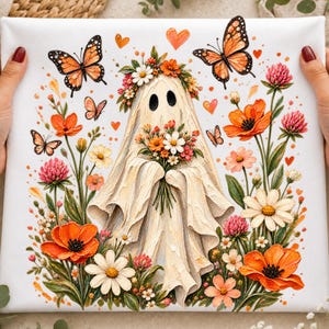 Può includere: Una stampa su tela quadrata raffigurante una figura di fantasma bianca centrale che indossa una corona di fiori e tiene in mano un bouquet. Il fantasma è circondato da farfalle monarca arancioni, vari fiori di campo e piccoli cuori arancioni.
