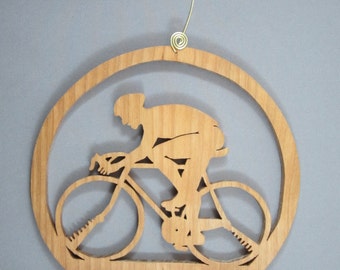 May include: A wooden ornament shaped like a circle with a cutout of a person riding a bicycle. The ornament has a gold wire hanger.