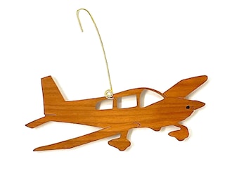 Könnte beinhalten: Ein Holz-Anhänger in Form eines kleinen Flugzeugs. Der Anhänger ist in hellbrauner Farbe bemalt und hat einen goldenen Aufhänger.