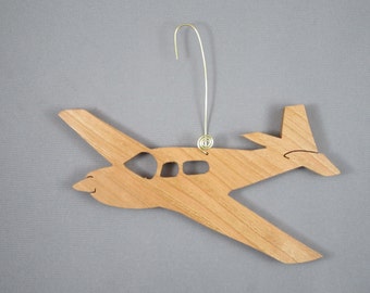 Könnte beinhalten: Holzflugzeug-Ornament mit natürlicher Holzoberfläche. Das Flugzeug hat ein detailliertes Design mit Flügeln, einem Heck und Fenstern. Ein goldfarbener Draht ist zum Aufhängen befestigt. Das Ornament ist ein dekorativer Artikel.