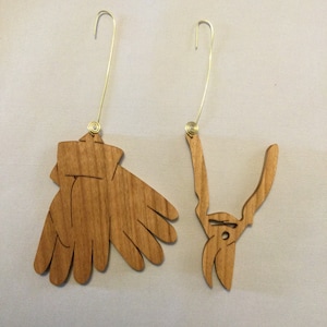 May include: Two wooden Christmas ornaments, one shaped like a pair of gardening gloves and the other shaped like a pair of pruning shears.