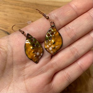 Può includere: Un paio di orecchini pendenti a forma di goccia con una finitura in smalto maculato nei toni del giallo dorato, verde oliva e bianco. I componenti metallici sono color rame e la superficie presenta piccoli ritagli circolari.