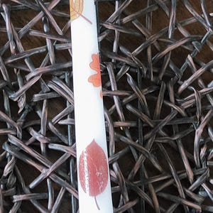 May include: A white taper candle featuring a repeating pattern of various autumn leaves in shades of orange, red, and brown. The candle has a smooth, cylindrical shape with a single white wick at the top, showcasing detailed leaf illustrations.
