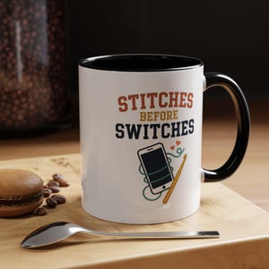 May include: A white ceramic mug with a black interior and handle. The front features the text STITCHES BEFORE SWITCHES in orange and dark blue, accompanied by a graphic of a smartphone wrapped in green yarn with a yellow crochet hook and hearts.