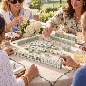 Puede incluir: Un juego de Mahjong con fichas de color verde p&aacute;lido que presentan s&iacute;mbolos, caracteres y dise&ntilde;os florales en negro y verde. Las fichas est&aacute;n dispuestas sobre un mantel claro, y algunas muestran las letras E, S, W, N y la palabra JOKER.