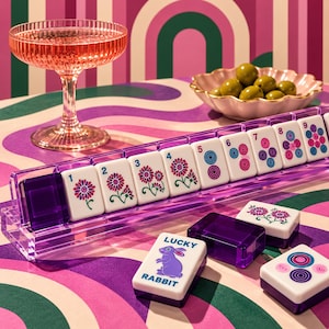 Puede incluir: Un juego de fichas de mahjong blancas con reverso púrpura translúcido, con diseños florales, números del uno al nueve y una ficha con un conejo púrpura y el texto LUCKY RABBIT. Las fichas están dispuestas en un soporte de acrílico púrpura.