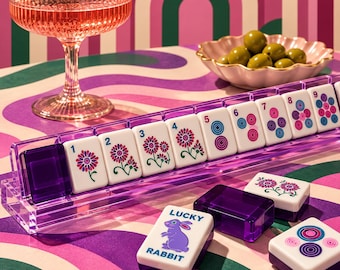 May include: A set of white mahjong tiles with purple translucent backs, featuring floral designs, numbers one through nine, and a tile with a purple rabbit graphic and the text LUCKY RABBIT. The tiles are displayed in a purple acrylic rack.