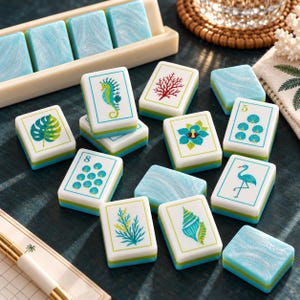 Puede incluir: Un juego de fichas de mahjong rectangulares con reverso jaspeado en azul y blanco y caras blancas con ilustraciones coloridas de vida marina, incluyendo un caballito de mar, coral, una flor de hibisco, un flamenco, conchas y algas, con algunos números.