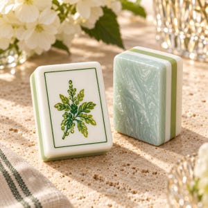 Puede incluir: Dos fichas de mahjong rectangulares con bordes redondeados. Una ficha presenta una cara blanca con una ilustraci&oacute;n bot&aacute;nica verde de una planta frondosa dentro de un borde verde fino. La otra ficha muestra un panel lateral verde claro nacarado.