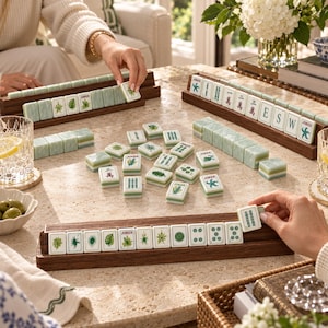 Puede incluir: Un juego de Mahjong con fichas de color verde claro con ilustraciones bot&aacute;nicas y num&eacute;ricas, dispuestas sobre soportes de madera oscura y una mesa de piedra. Las fichas muestran motivos vegetales, caracteres chinos y la palabra Joker.