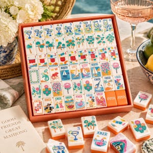 Puede incluir: Un juego de mahjong con fichas blancas que presentan ilustraciones coloridas de cócteles, palmeras, veleros, cactus y flores. Las fichas están dispuestas en una bandeja de madera, con otras sueltas delante, junto a un libro titulado Good Friends Great Mahjong.
