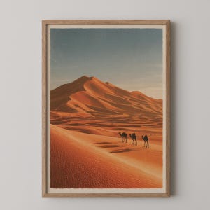 May include: A framed wall art print featuring a desert landscape with large orange sand dunes under a clear sky. Three camels are walking in a line across the sand. The artwork is enclosed in a light-colored wooden frame with a visible grain.
