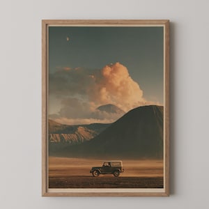 Op de afbeelding: Een ingelijste wandkunstprint met een landschapsscène waarin een donker terreinvoertuig over een uitgestrekte, stoffige vlakte rijdt. Op de achtergrond verrijzen grote bergen onder een lucht vol goudkleurige wolken en een kleine maan.